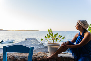 Beautiful woman with blue sarong and Greek outfit on her head relaxingly enjoying the spectacular view of the sea to the south of the greek island of Crete in summer from a Greek tavern