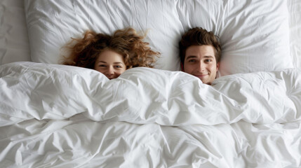 A couple peeks out playfully from under a white duvet in bed, their expressions full of joy and comfort in an intimate and cozy bedroom setting.