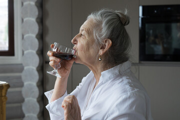 a woman of retirement age in a white blouse drinks wine by the fireplace
