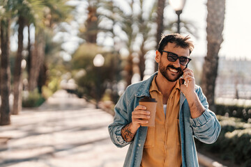 A young fashionable guy is talking on his phone on a city street and holding coffee to go.