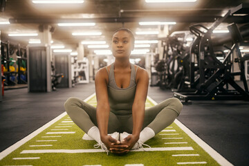 A dedicated fit black sportswoman is sitting on gym floor and stretching her legs.