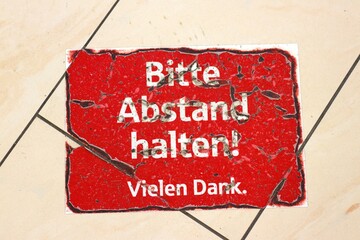 Worn-out red sign on tiled floor in German: Bitte Abstand halten. Vielen Dank. Translated to:...
