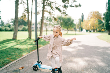 Little girl stands leaning on a scooter on the road in the park