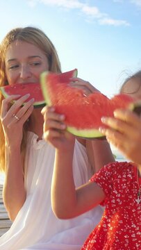 Happy mother and her daughter sitting on a wooden pier and eating a juicy red watermelon in the sunshine.