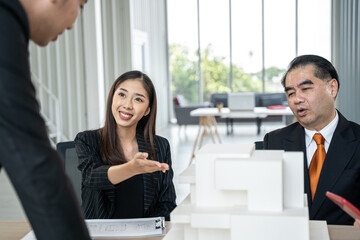 An animated businesswoman gestures during a discussion with male colleagues over model building plans in a light-filled corporate office.