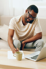 Smiling African American man working on laptop while sitting on a modern sofa in a cozy indoor...