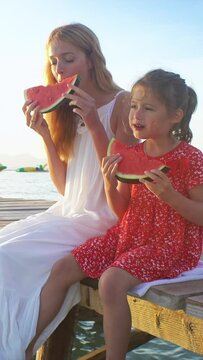 Happy mother and her daughter sitting on a wooden pier and eating a juicy red watermelon in the sunshine.
