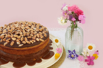 Homemade peanut cake and caramel sauce decorated with peanuts on pink background with summer flowers. Delicious afternoon tea floral composition.