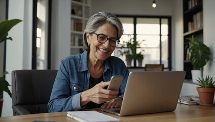 Smiling mature Hispanic woman engaged in a video call on her smartphone while working with a laptop in a bright home office Mother's day Women's day