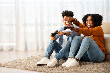 Joyful African American couple playing video games on the floor, with the woman playfully covering...