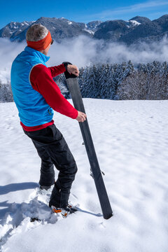 Ski waxing.  Man stands in the snow and holds a ski in his hand.  He waxes the ski with his other hand.  Mountainous winter landscape, sunny day, blue sky.