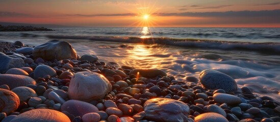 The beach is covered in numerous rocks of varying sizes near the waters edge as the sun sets, casting a warm glow over the scene. Waves gently splash against the rocks, creating a serene atmosphere.