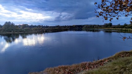 A mixed forest grows on the grassy banks of the river. The river is making a turn. In the fall, the leaves on the trees turn yellow. There are ripples on the water and the reflection of the sunset