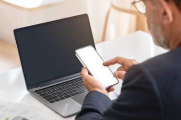 A businessman in a suit uses a smartphone with a blank screen alongside an open laptop