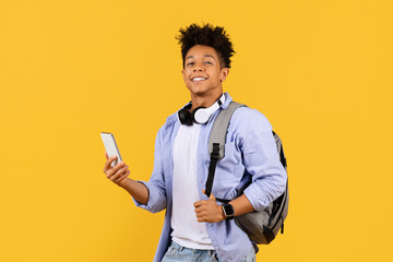 Joyful black male student with headphones and phone on yellow background