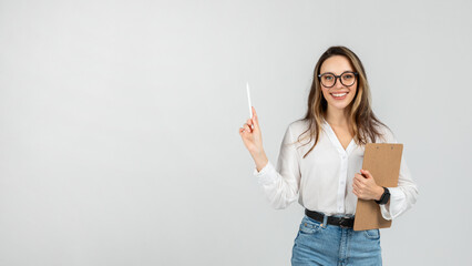 A smiling young woman with glasses holding a clipboard and pointing up with a pen