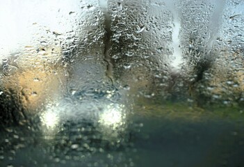 Abstraktes Motiv mit Wassertropfenmuster auf beschlagener nasser Glasscheibe vor weißem Auto mit weißen Lichtern auf Straße in Stadt bei Frost, Kälte und Regen am Morgen im Winter