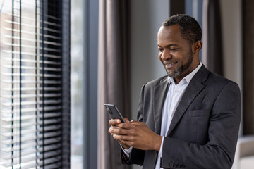 Smiling african american businessman in suit using smartphone by window
