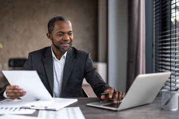 Professional african american businessman working at office desk with laptop and documents - obrazy, fototapety, plakaty
