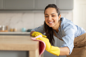 Happy young woman dusting off wooden table at kitchen