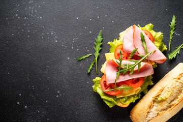 Ciabatta sandwich with lettuce, cheese, tomatoes and ham on black table.