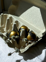 Black and gold eggs lie in a tray on a light background under the rays of the sun, next to a bunch of willow. Celebration of the bright Easter holiday. Vertical photo