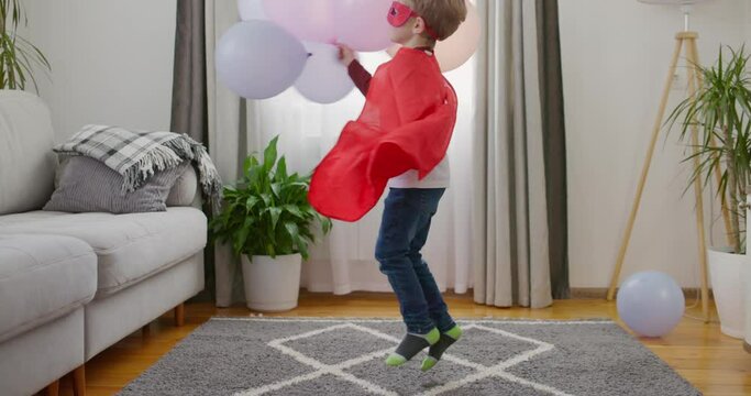 Child in superhero costume with red cape and mask playing with balloons in home
