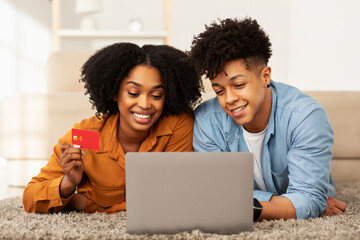 Happy African American couple lying on the floor with a laptop, woman holding a credit card