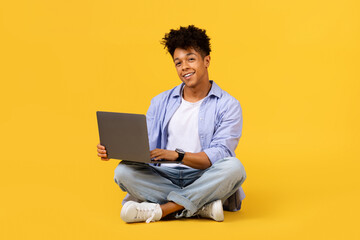 Smiling black student guy with laptop sitting cross-legged on yellow background