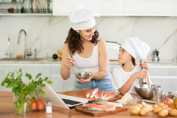 Happy mother and daughter are baking salmon in the kitchen while looking at the recipe on laptop
