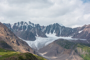 Dramatic landscape with glacier tongue among rocky ridges and sharp snow-capped mountain range under gray cloudy sky. Alpine valley among green hills and rocks in sunlight against ice and sheer crags.