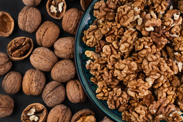 On a dark background, peeled walnuts in a plate and unshelled ones on a dark table.