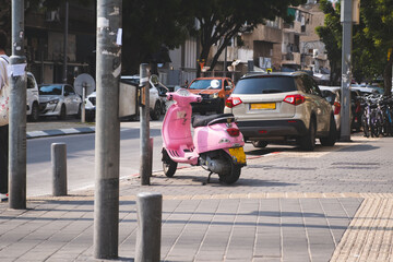 Fototapeta premium Pink scooter parked on a sunny street in Tel Aviv, Israel, with cars and bicycles in the background.