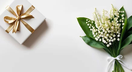  Un bouquet de muguet pour la fête du travail le 1er Mai et un cadeau blanc décoré avec un ruban doré sur un fond blanc © infographiste06