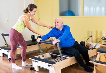 Young woman professional pilates instructor helping aged man doing exercises on reformer. Wellness concept for elderly