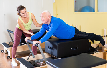 Focused positive senior man practicing pilates system on reformer to improve and maintain mobility under supervision of qualified female trainer