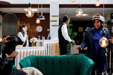 Naklejka premium Asian girlfriend with wintersports equipment excitingly joins boyfriend on cozy sofa in luxury winter resort. Happy woman wearing winter jacket and ski helmet approaches her friend in lounge area.