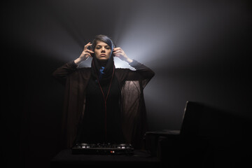 Woman holding headphones and operating DJ mixer inside a dark room.