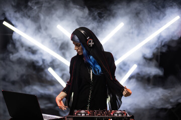 A female DJ with vibrant blue hair is actively mixing music at a turntable during a live set. Smoke...
