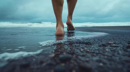 Foto op Canvas Closeup low angle shot of the adult male person or man walking barefoot on the dark gray sand beach, ocean or sea waves crushing under his feet or legs © Nemanja