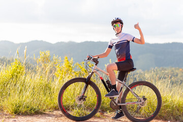 Brazilian cyclist standing with his bike and giving a thumbs up sign with his hand. He is happy and smiling while looking at the camera. In the background, landscapes with hills.