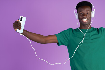 Happy young African man listening to music and dancing with headphones connected to his phone with a lilac background.