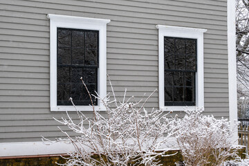 The exterior wall of a grey colored vintage building with narrow wooden clapboard siding, two double hung windows, black spacers, white decorative wood trim and a shrub covered with fresh white snow. 
