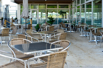 An outdoor eating area with chrome square tables and rattan chairs. The large open space is empty....