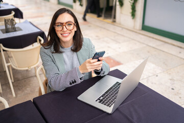 Brunette woman in casual clothes working with laptop using mobile phone at cafe. Freelancer using...