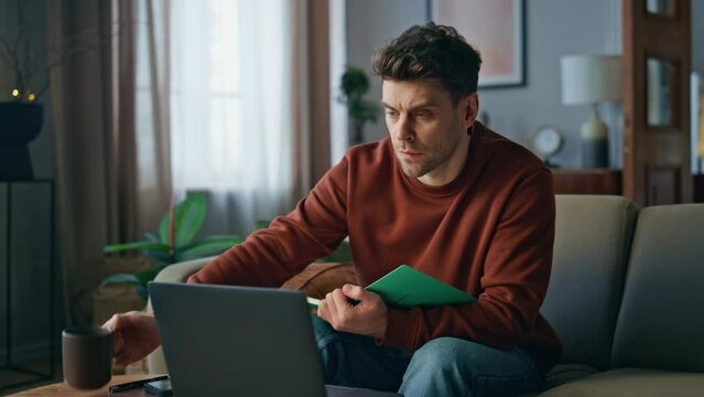 Guy watching online course at laptop sitting couch. Man drinking coffee studying