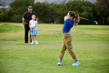 Obraz premium A father and daughter watch as other daughter swings her club on a fairway of a golf course