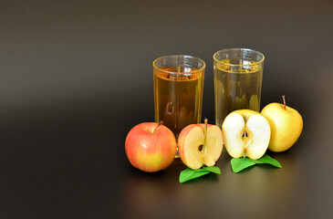 Two glass transparent glasses with different fruit juices on a black background, next to each halves of ripe red and yellow apples.