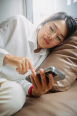 Side view of asian Thai office woman wear glasses using mobile phone while leaning and resting on brown couch sofa, tired of work. 