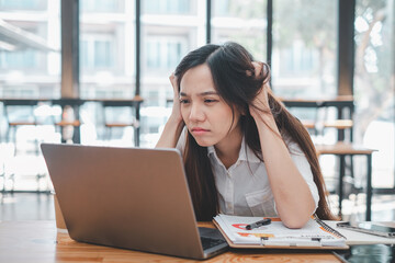 A young woman looks frustrated and stressed while concentrating on her laptop screen in a bustling cafe environment.